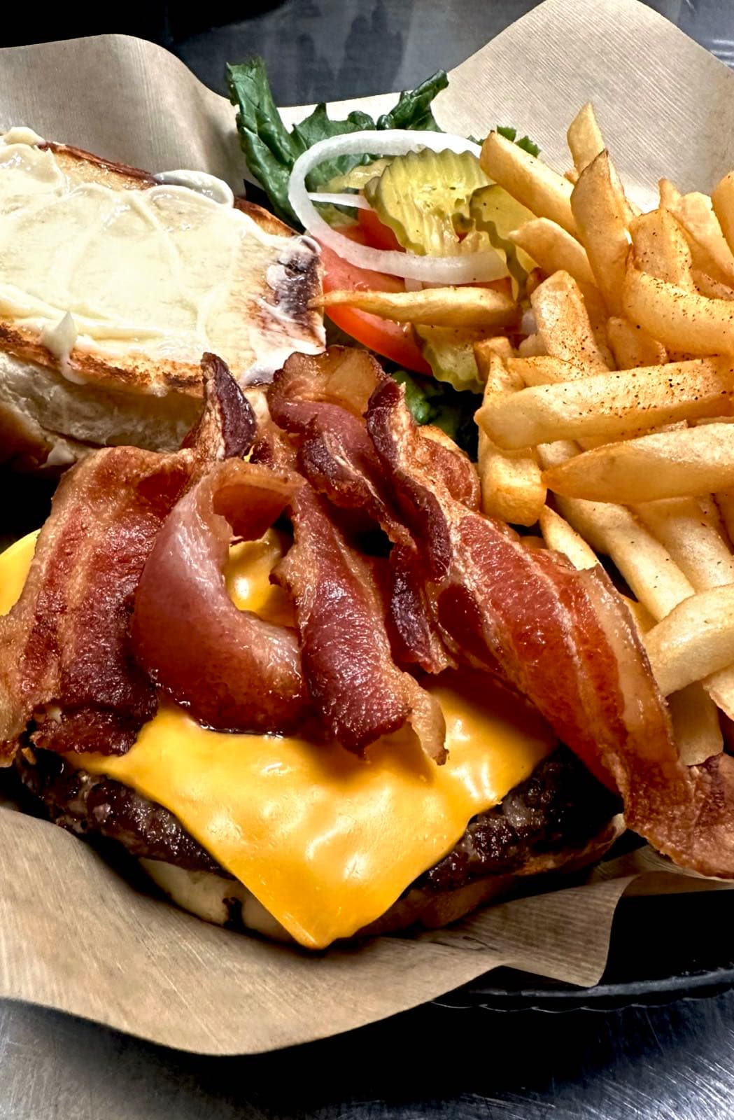Close-up of a bacon cheeseburger piled high with crispy bacon, melted cheese, lettuce, tomato, onion, and pickle, served with seasoned fries.