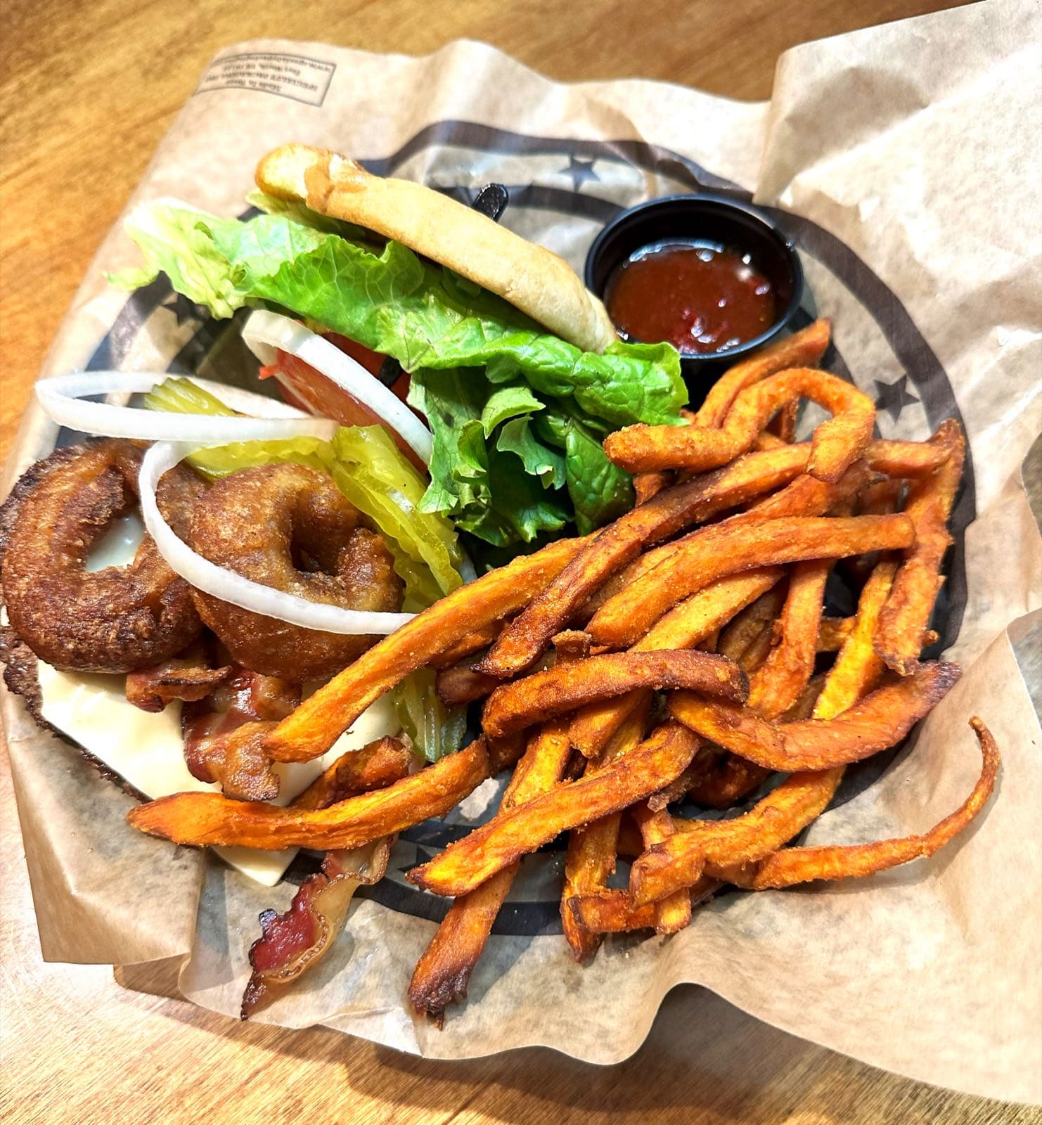 Overhead view of a bacon cheeseburger topped with onion rings, lettuce, tomato, onion, and pickles, served with a side of seasoned sweet potato fries.