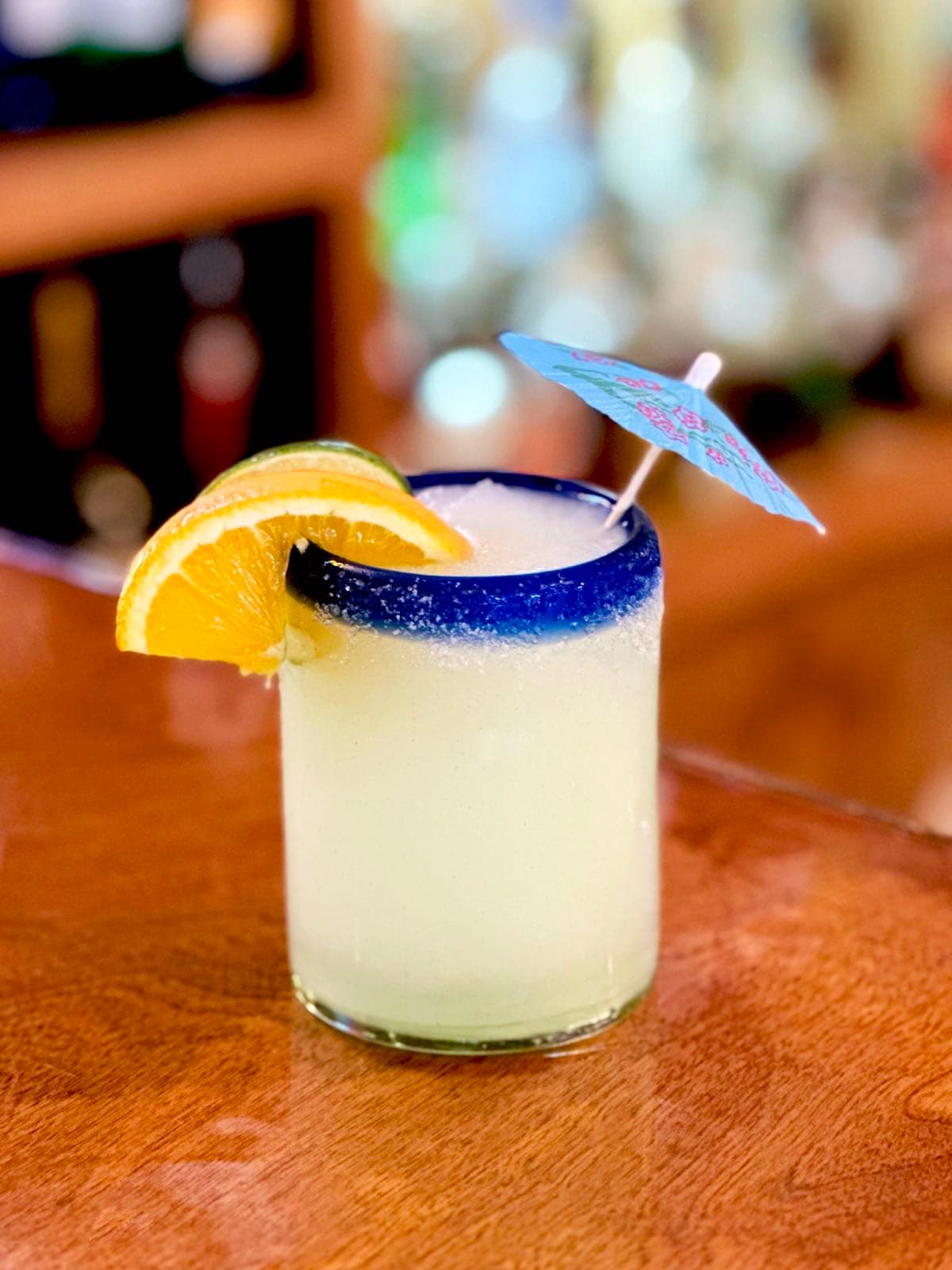 Salt-rimmed margarita with a blue-painted glass rim, garnished with an orange slice, lime wheel, and a paper cocktail umbrella.