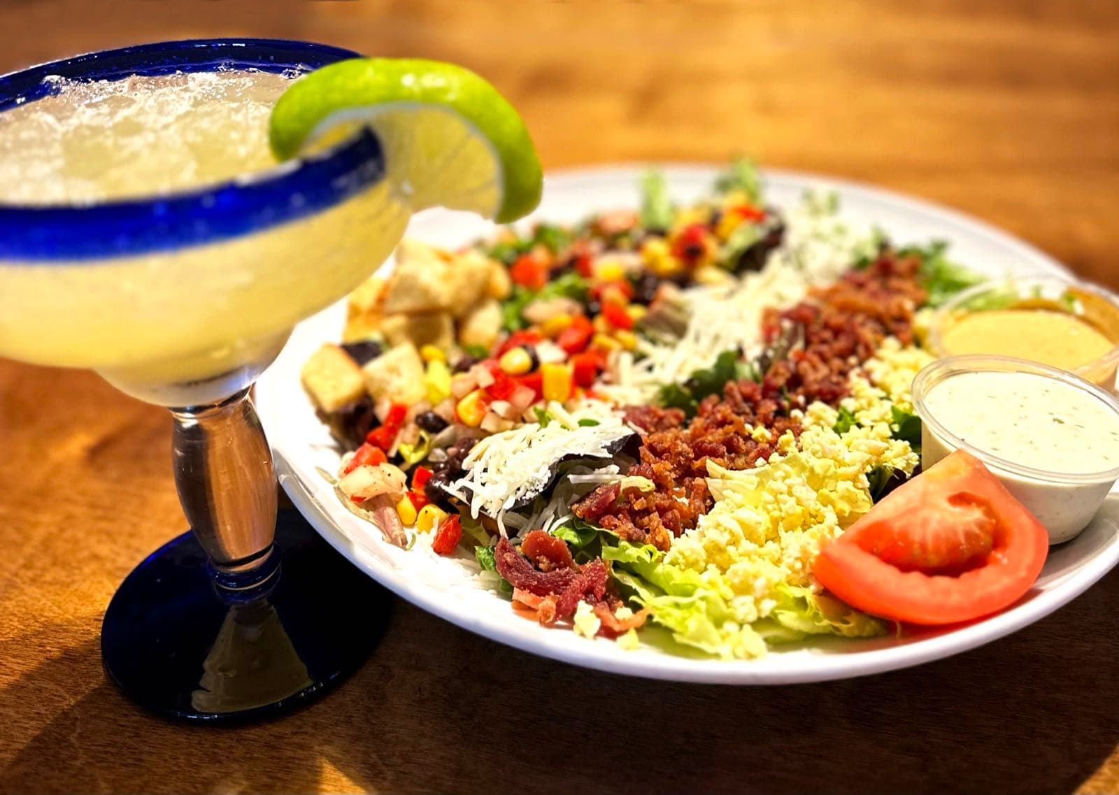 A salt-rimmed margarita with a lime wheel garnish next to a chopped salad topped with black beans, corn, pico de gallo, cheese, bacon, egg, and a tomato wedge.