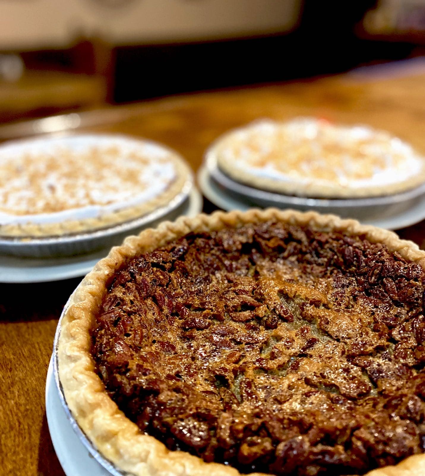 Three freshly baked pies on a wooden table, with a glossy pecan pie in sharp focus in the foreground.