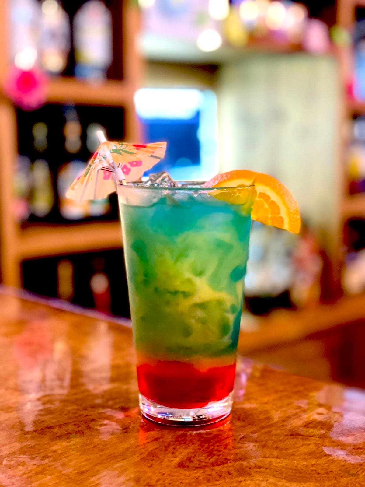 A tall layered cocktail in red, green, and blue, garnished with an orange slice and a paper umbrella, served on the bar at Texas Traditions.