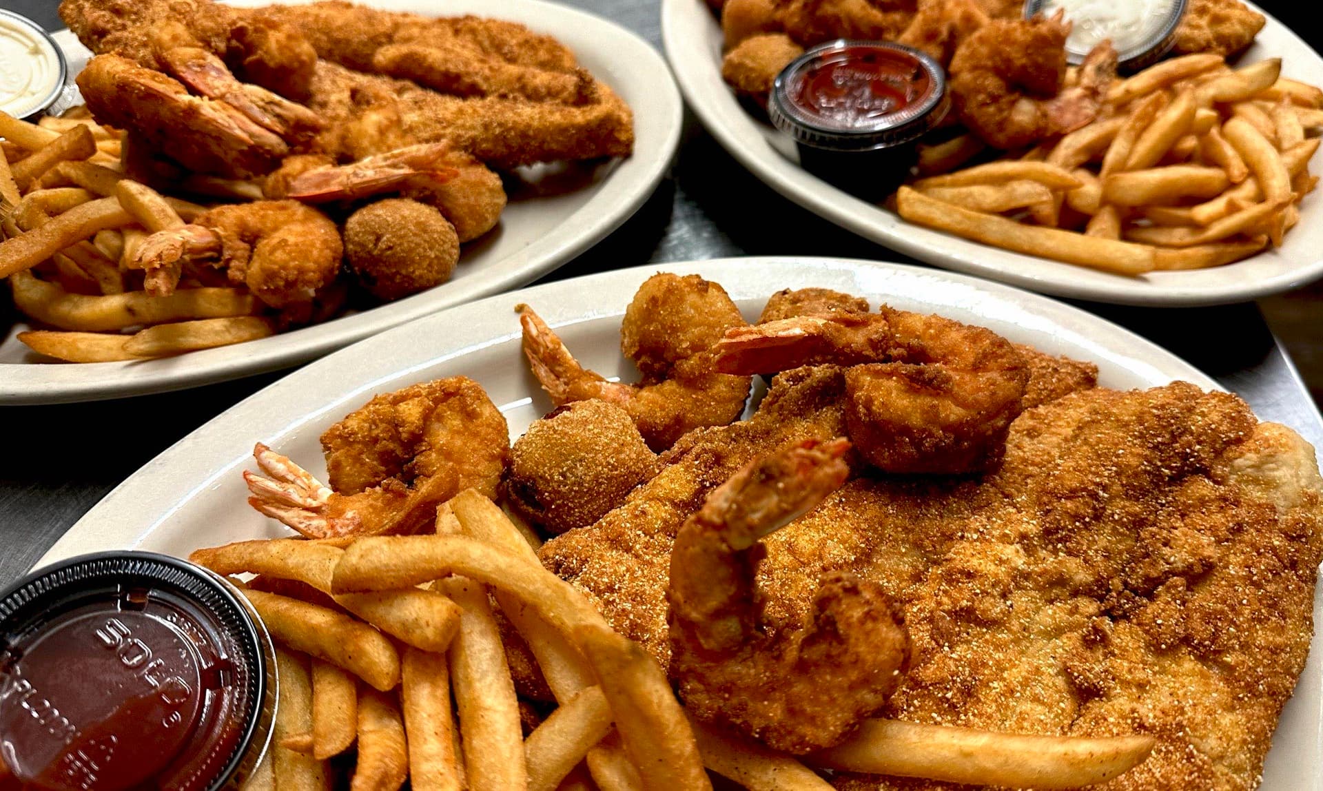 Three oval plates loaded with golden fried fish fillets, fried shrimp, hush puppies, and French fries, served with cocktail and tartar sauce.